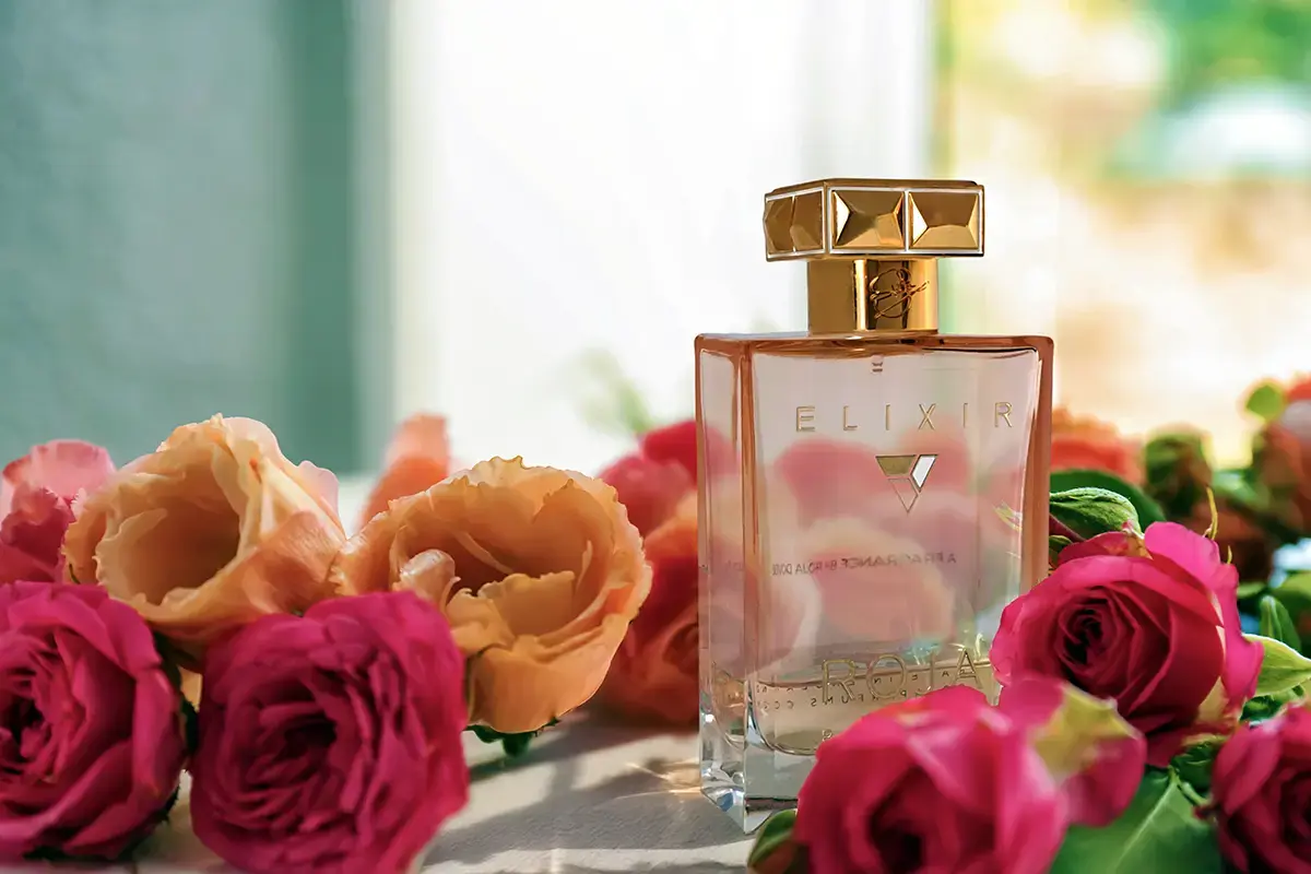 Roja Elixir Pour Femme surrounded by vibrant orange and pink roses, basking in sunlight